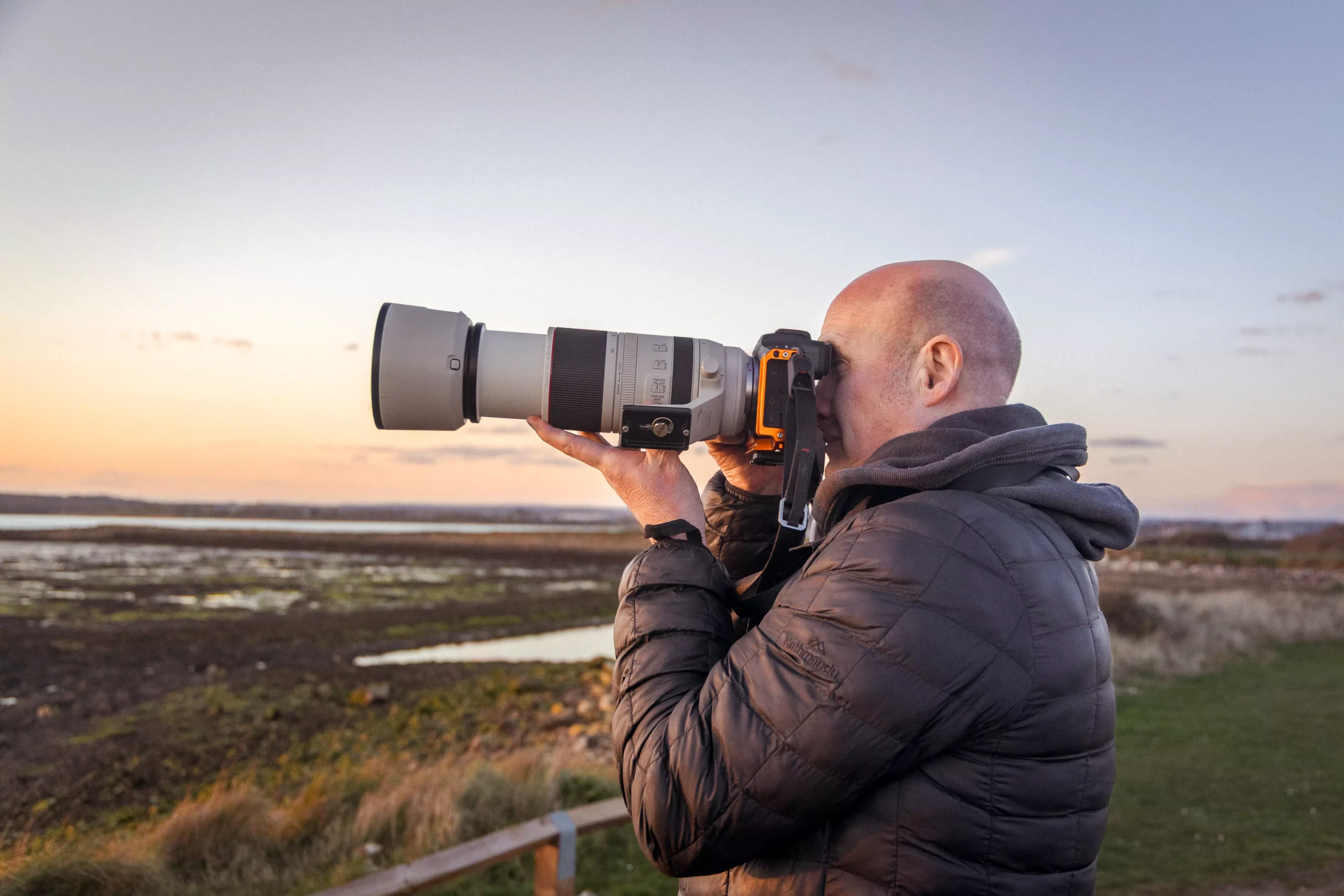 landscape photographer standing in a coastal forest, holding a camera and tripod, wearing a dark jacket