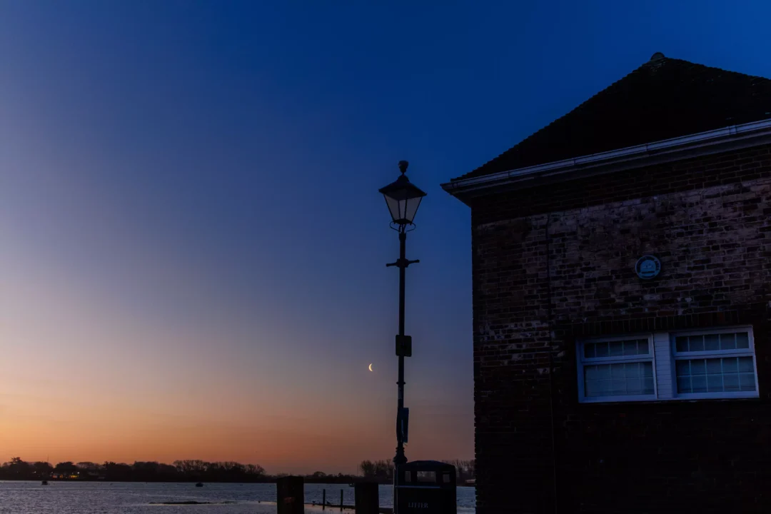 A thin crescent moon hangs in a deep blue twilight sky next to a traditional street lamp and a brick waterfront building.