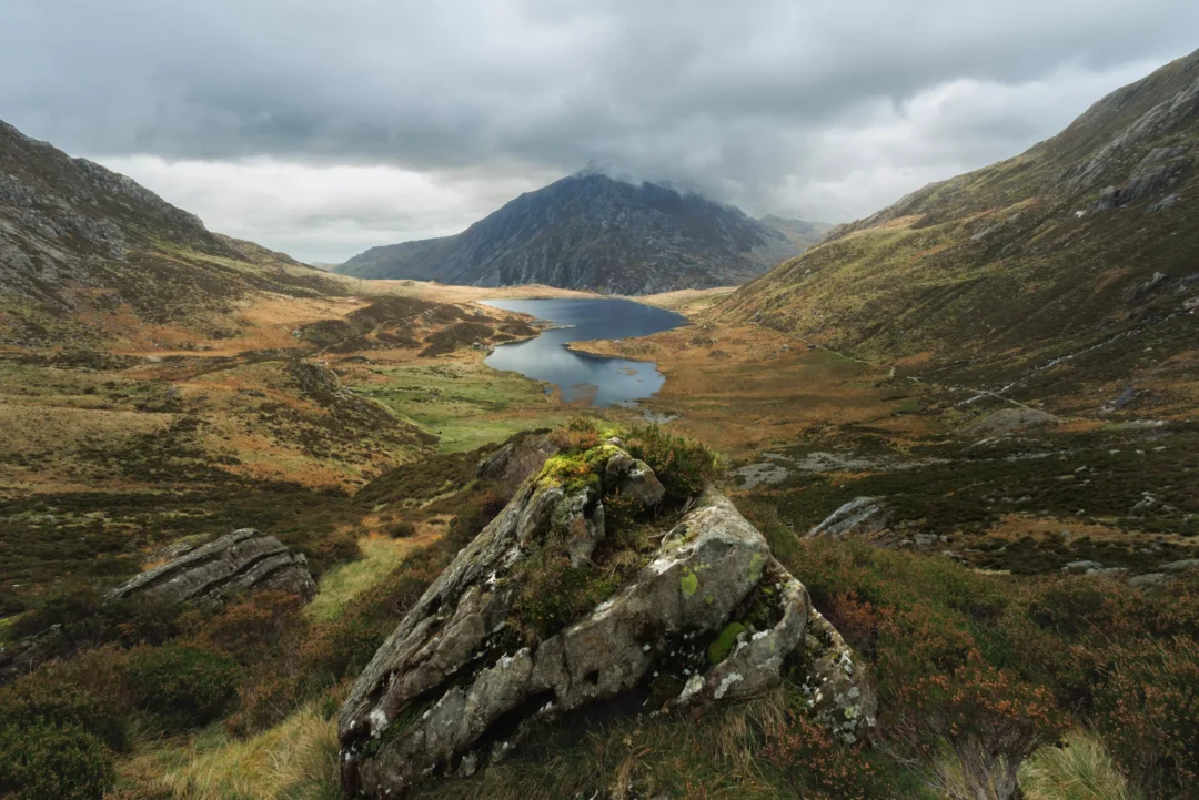 An elevated view looking down into Cwm Idwal, Snowdonia, with Llyn Idwal visible far below, the valley floor in rich autumn colour and Pen yr Ole Wen rising beyond