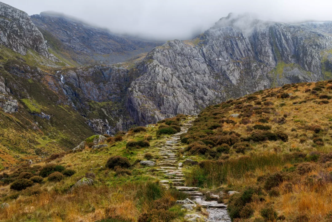 A stone-paved path climbing through open moorland towards the cliffs of Cwm Idwal, Snowdonia, with waterfalls visible on the distant rock face and low cloud on the summits