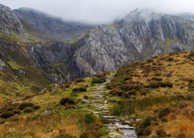 A stone-paved path climbing through open moorland toward the cliffs of Cwm Idwal, Snowdonia, with waterfalls visible on the distant rock face and low cloud on the summits