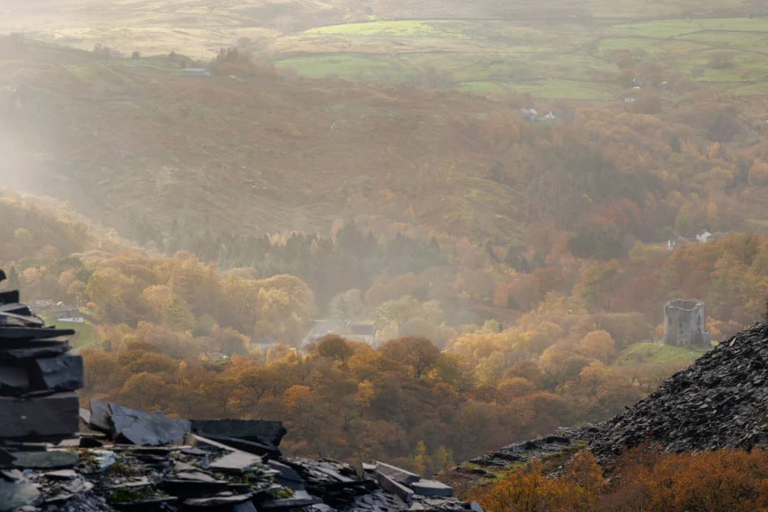 A distant view of Dolbadarn Castle tower sitting on a rocky outcrop surrounded by misty autumn woods and Llyn Peris.
