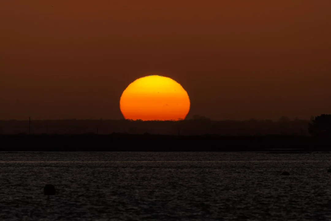 A perfectly round yellow and orange sun rising directly over a dark landmass and rippling coastal waters.