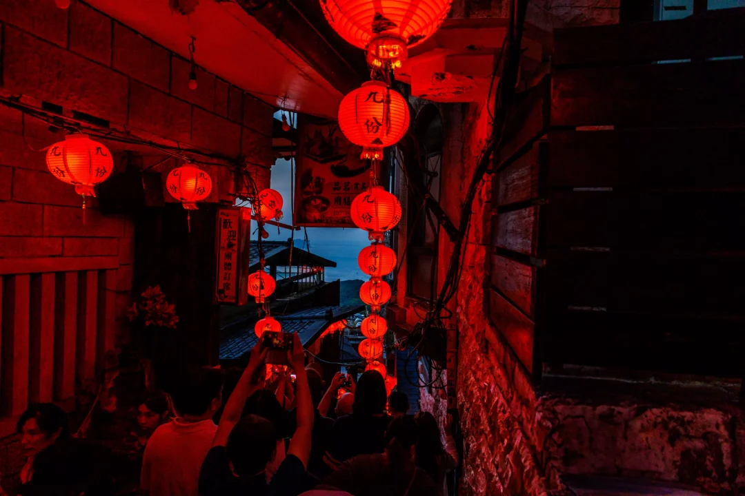Narrow Jiufen alleyway bathed in deep red lantern light at night with crowds and the dark sea visible at the end