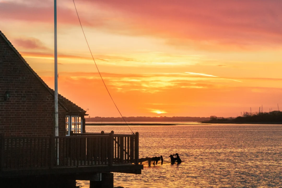 Brick mill building and wooden jetty silhouette against a fiery orange and pink dawn sky over the water.