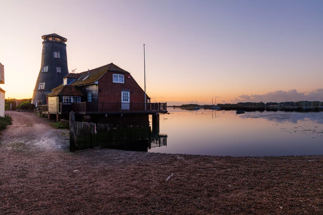 The iconic dark windmill tower and brick mill building at the water’s edge during a calm, clear dawn.