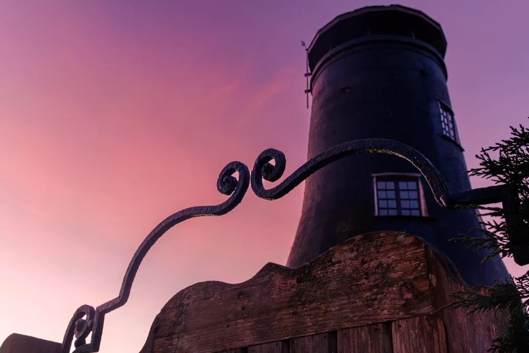 A close-up of a weathered wooden gate with decorative iron scrolls, with the dark silhouette of Langstone Windmill against a purple dawn sky.