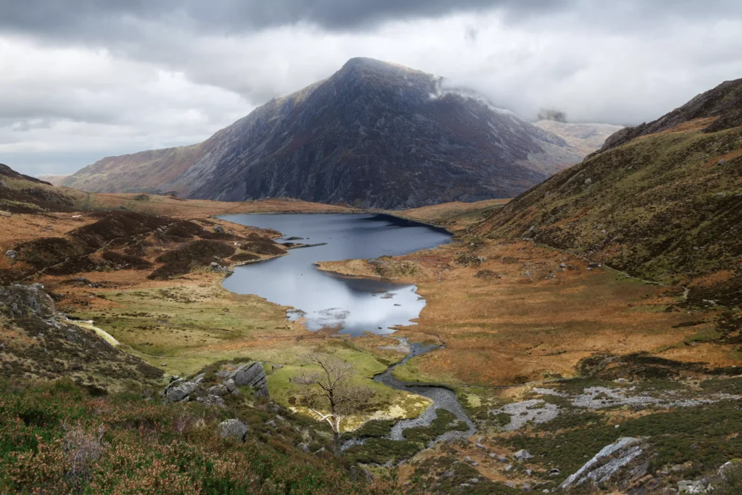 Llyn Idwal reflecting the headwall cliffs of Cwm Idwal, Snowdonia, with a waterfall visible on the far cliff face and golden autumn grasses on the shoreline