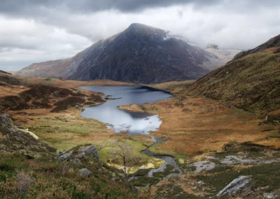 Llyn Idwal reflecting the headwall cliffs of Cwm Idwal, Snowdonia, with a waterfall visible on the far cliff face and golden autumn grasses on the shoreline