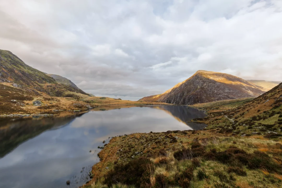 Llyn Ogwen reflecting Pen yr Ole Wen mountain under dramatic clouds, with warm golden light on the hillside and a wide sky over the Ogwen Valley, Snowdonia