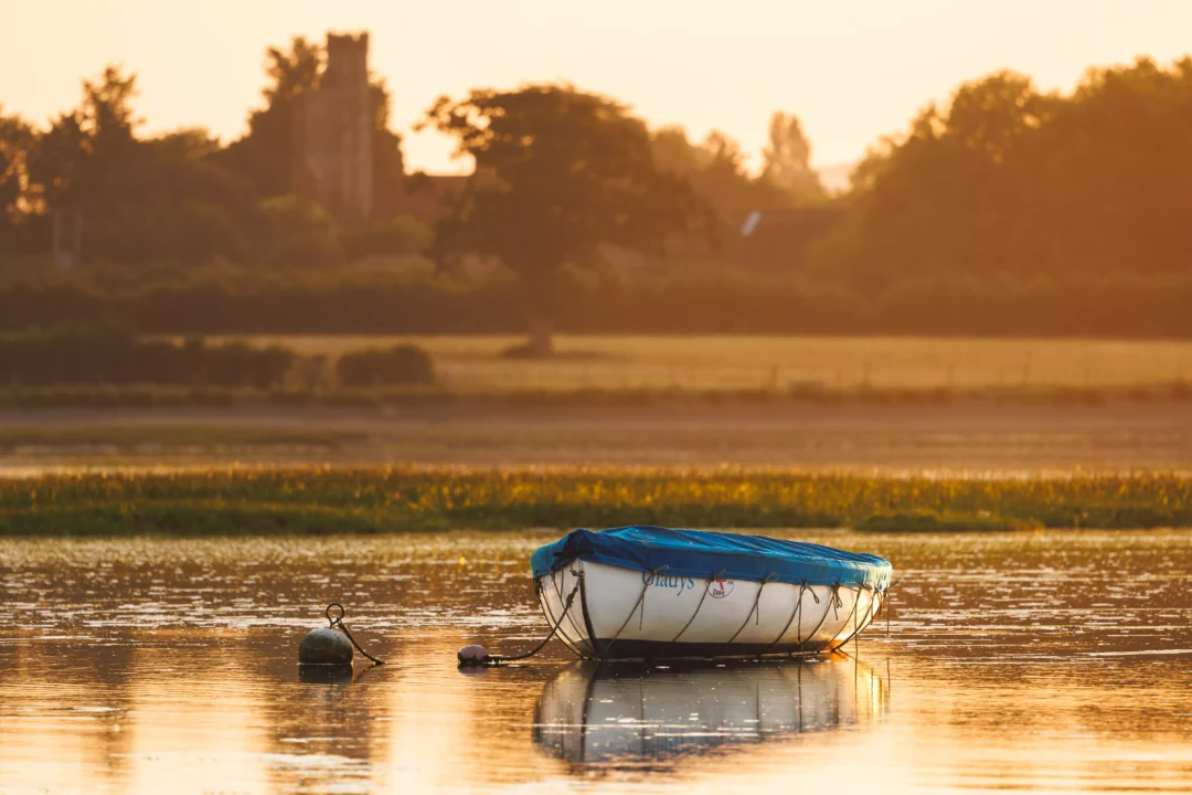 A white dinghy with a blue cover resting on calm water with a soft-focus historic stone tower in the distance.