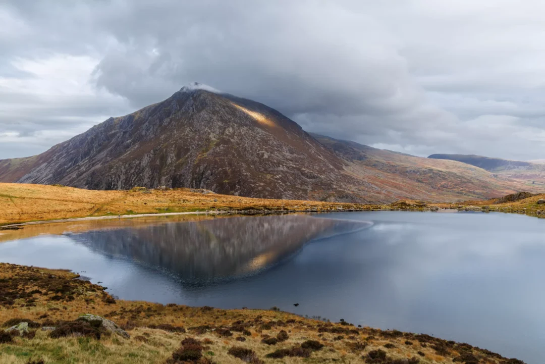 Pen yr Ole Wen reflected in the still waters of Llyn Ogwen, Ogwen Valley, Snowdonia, with dramatic storm clouds and a patch of golden light on the summit ridge