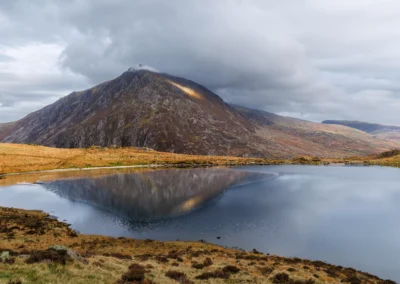 Pen yr Ole Wen reflected in the still waters of Llyn Ogwen, Ogwen Valley, Snowdonia, with dramatic storm clouds and a patch of golden light on the summit ridge
