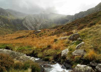 A rainbow over the Ogwen Valley, Snowdonia, with Llyn Ogwen visible in the middle distance and golden moorland grasses in the foreground under a grey sky