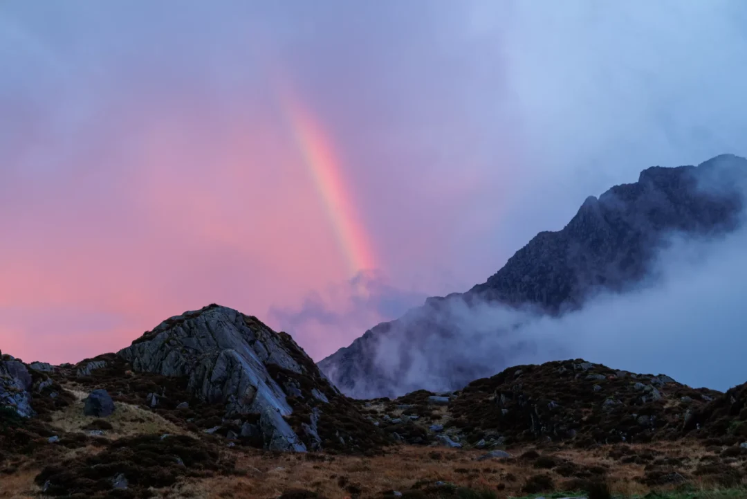 A vivid rainbow arcing through a pink and purple dusk sky above Tryfan, Ogwen Valley, Snowdonia, with mist rolling across the mountain's rocky face