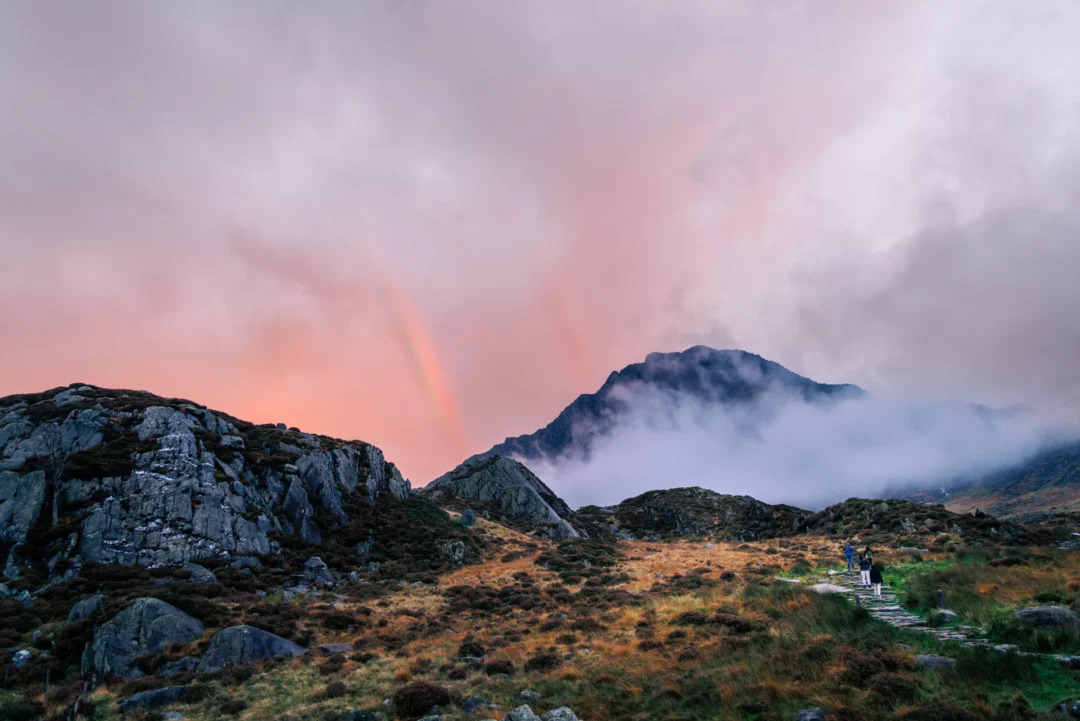 A mountain waterfall cascading over rocks at sunset in the Ogwen Valley, Snowdonia, with a burning orange sky and a bare twisted tree silhouetted against the clouds