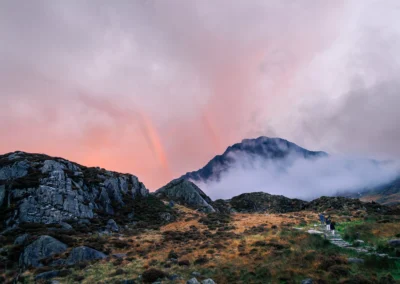 A mountain waterfall cascading over rocks at sunset in the Ogwen Valley, Snowdonia, with a burning orange sky and a bare twisted tree silhouetted against the clouds