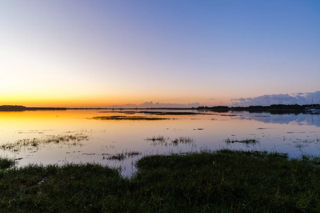 A wide view of the coastal marsh and still water reflecting a gradient of yellow to deep blue early morning sky.