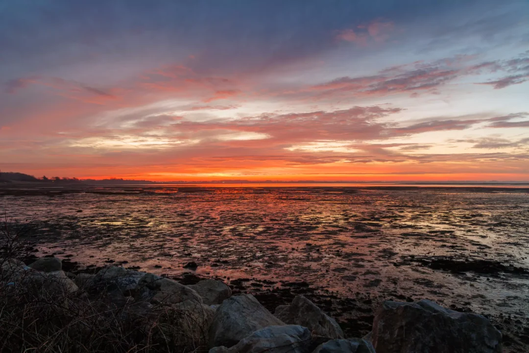 Large foreground rocks on a coastal bank looking out across a wide bay at a soft orange and grey sunrise.