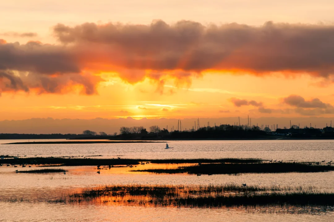 Sunbeams breaking through dark, low-hanging orange clouds over a quiet coastal tide with distant boat masts.