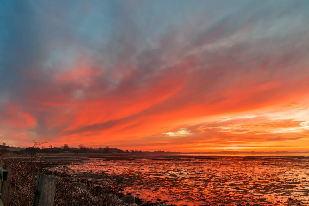 A dramatic morning sky filled with fiery orange and red clouds reflecting intensely across wet, dark mudflats.