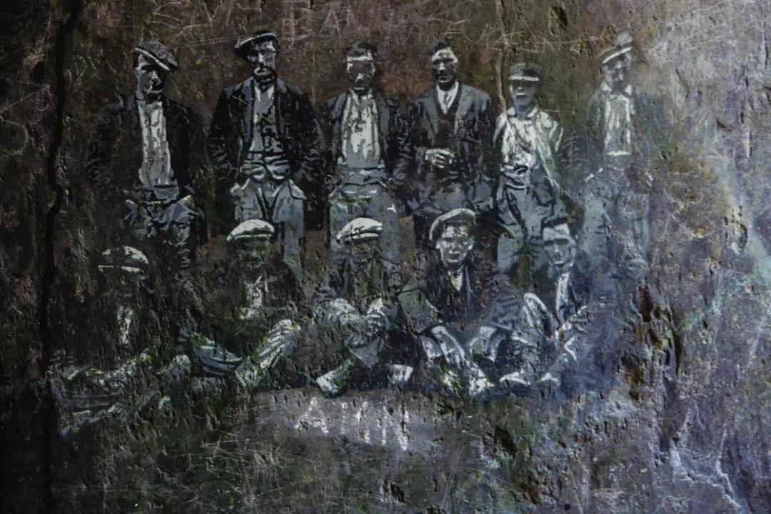 A detailed white-line engraving on a dark slate slab depicting a bearded quarry worker with a moustache and hat.