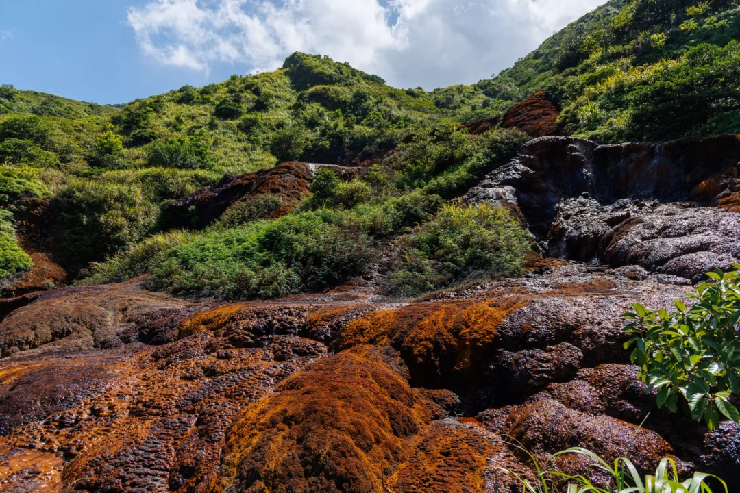 Wide view of Xiaoyoukeng volcanic area with red mineral rock formations and a small waterfall among green vegetation