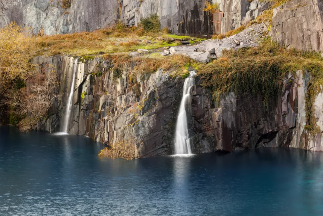 Two waterfalls spilling off a mossy slate ledge into a flooded blue quarry pit at Dinorwig.