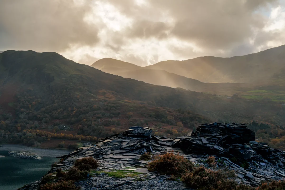 Dramatic sunbeams breaking through dark clouds over the mountains and Llyn Padarn lake, seen from a slate waste pile.