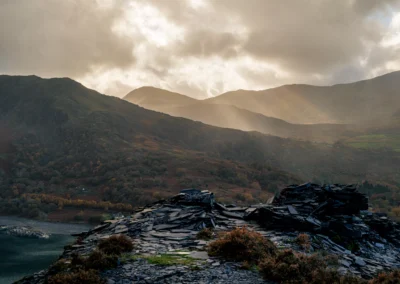 Dramatic sunbeams breaking through dark clouds over the mountains and Llyn Padarn lake, seen from a slate waste pile.
