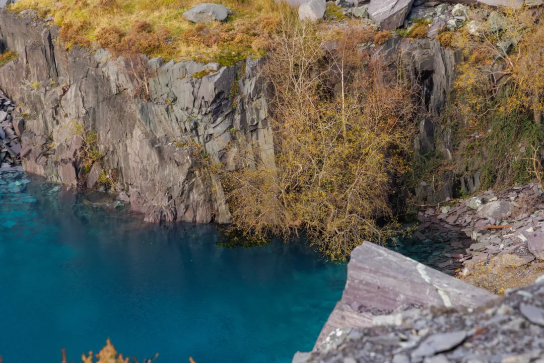 A narrow white waterfall cascading down a tiered slate cliff into a bright turquoise blue quarry pool.