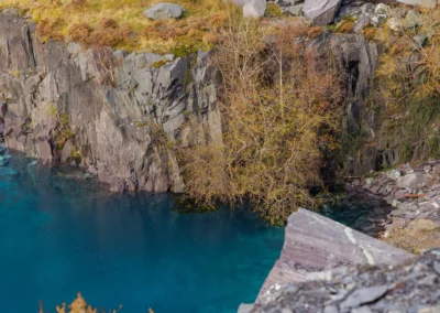 A narrow white waterfall cascading down a tiered slate cliff into a bright turquoise blue quarry pool.