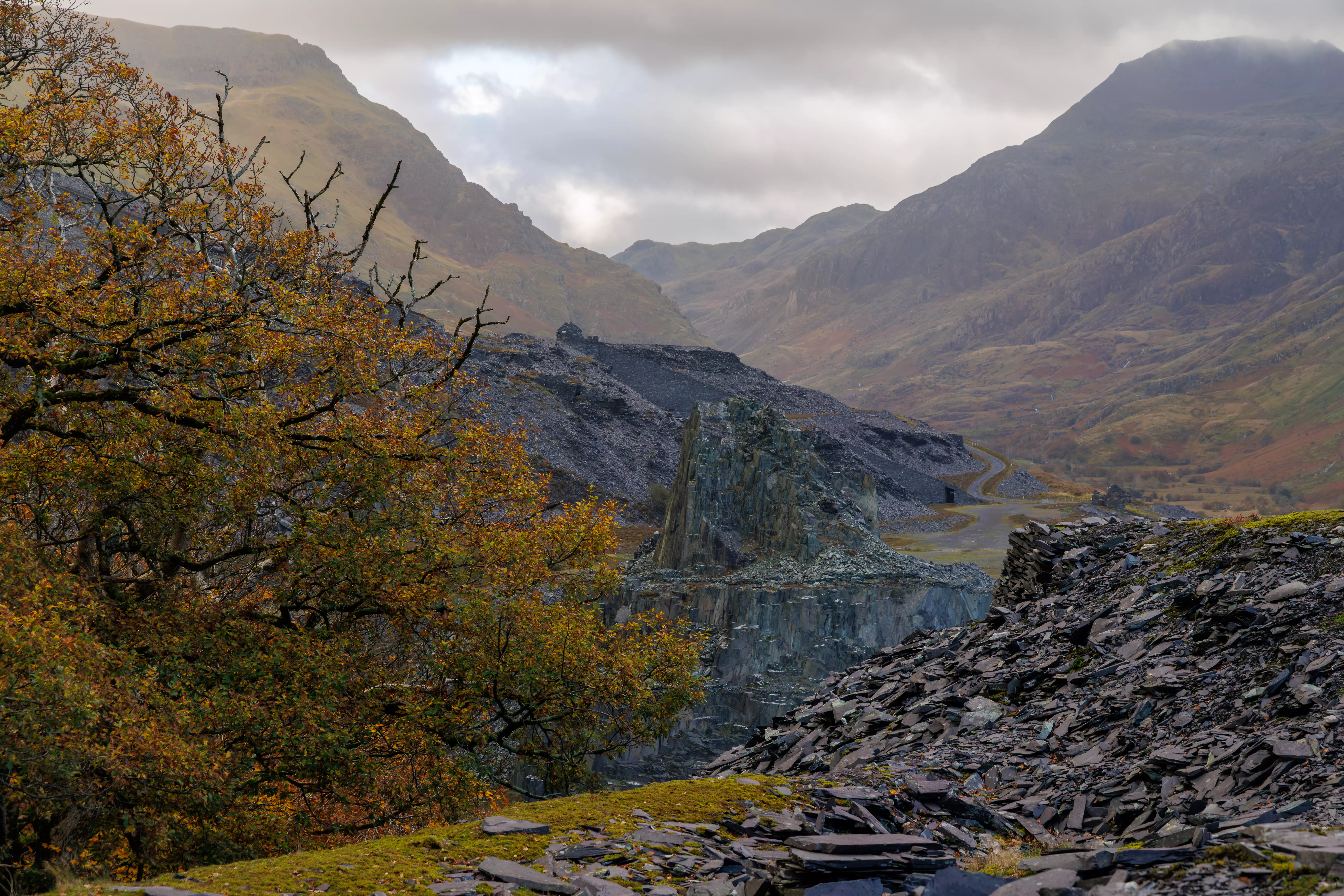 A high-angle landscape view looking down into the Llanberis Pass valley with a road and lake visible through the haze.