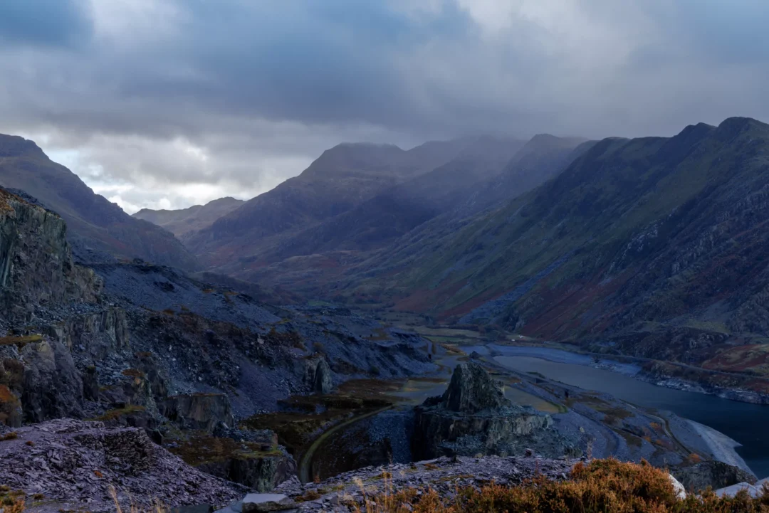 A moody, low-light landscape showing the dark silhouettes of mountains flanking Llyn Peris with soft light on the water.