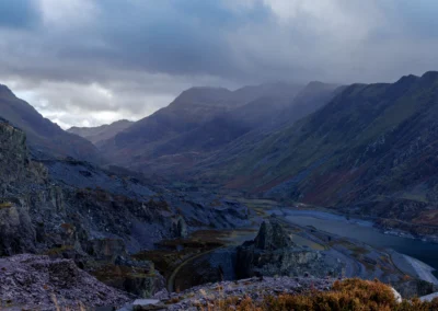 A moody, low-light landscape showing the dark silhouettes of mountains flanking Llyn Peris with soft light on the water.