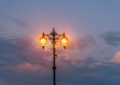 An intricate wrought-iron streetlamp glows with a warm amber light against a deep violet and pink stormy twilight sky.