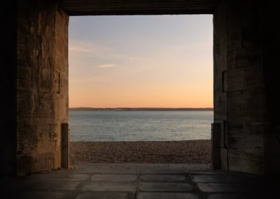 Heavy, textured stone walls frame a rectangular view of a pebble beach and the calm, pale orange horizon of the Solent at dusk.