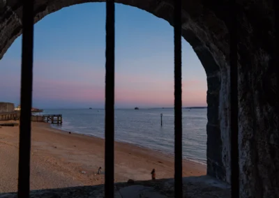 A high-angle coastal view of the shingle shore and soft pink evening sky, framed by the dark iron grille of a historic stone fortification.