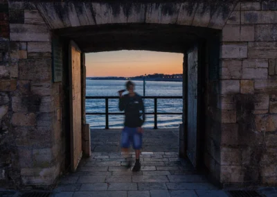 Looking through a heavy stone archway towards the sea at dusk, with a blurred silhouette of a person passing through.