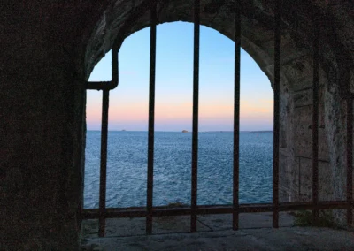A view of the blue sea and sunset sky framed through the dark iron bars of an old stone embrasure at the Hot Walls.