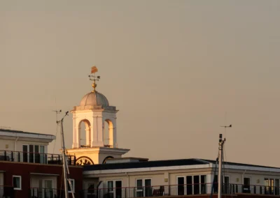 Warm golden hour light strikes a classic white wooden cupola and clock tower standing proud above coastal residential buildings.