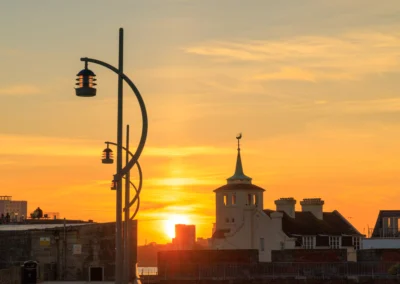 Stylised curved streetlamps stand silhouetted against a brilliant yellow and orange sky above the historic coastal defensive walls.