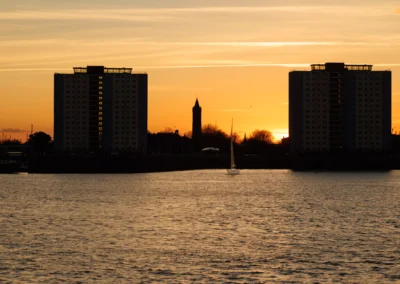 A lone white yacht sails across the glowing orange water between two large apartment buildings silhouetted against the setting sun.