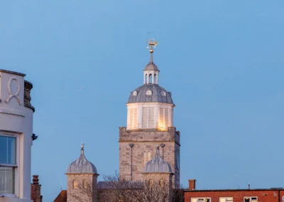 The stone tower and illuminated white cupola of the historic church topped with a golden ship weather vane against a clear blue sky.