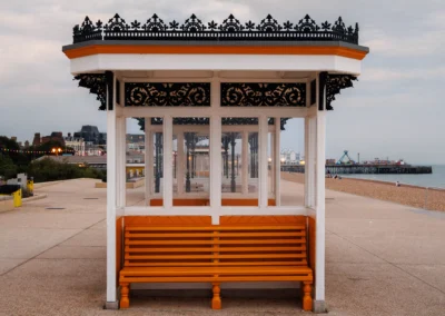 A clean, symmetrically framed white and orange shelter with decorative ironwork, looking down the beach towards Clarence Pier.