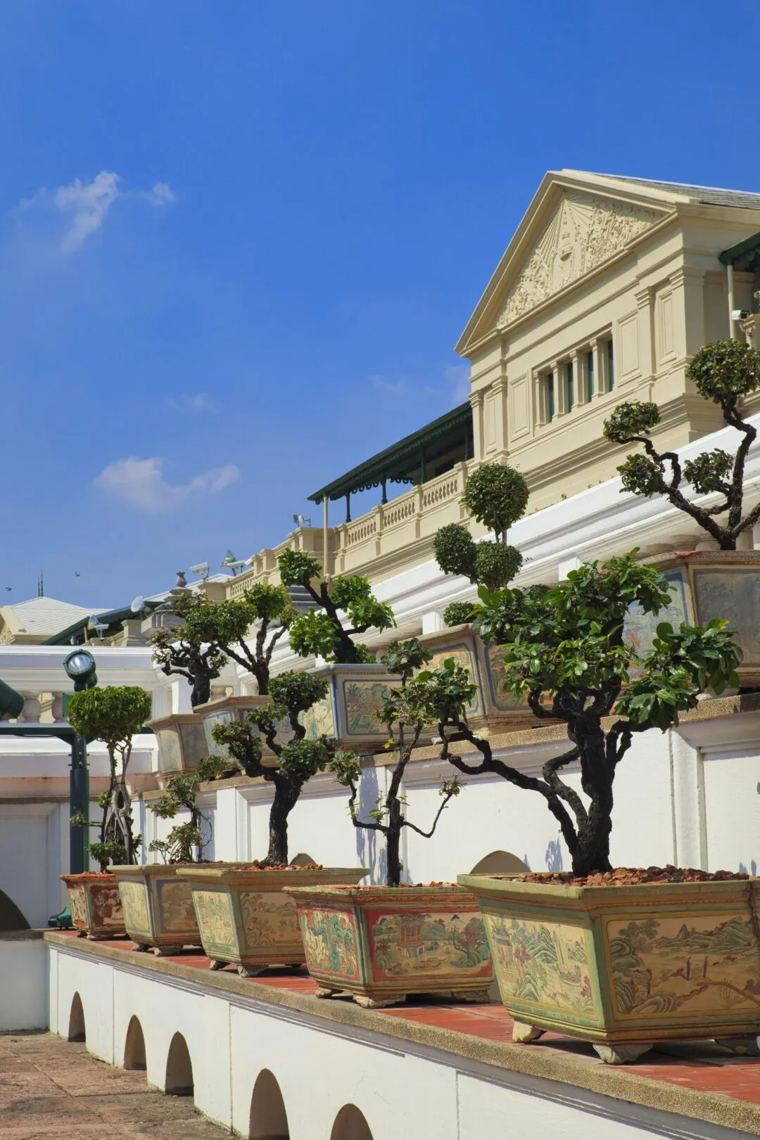 A row of sculpted bonsai trees in painted pots along a colonial-style wall in Bangkok.