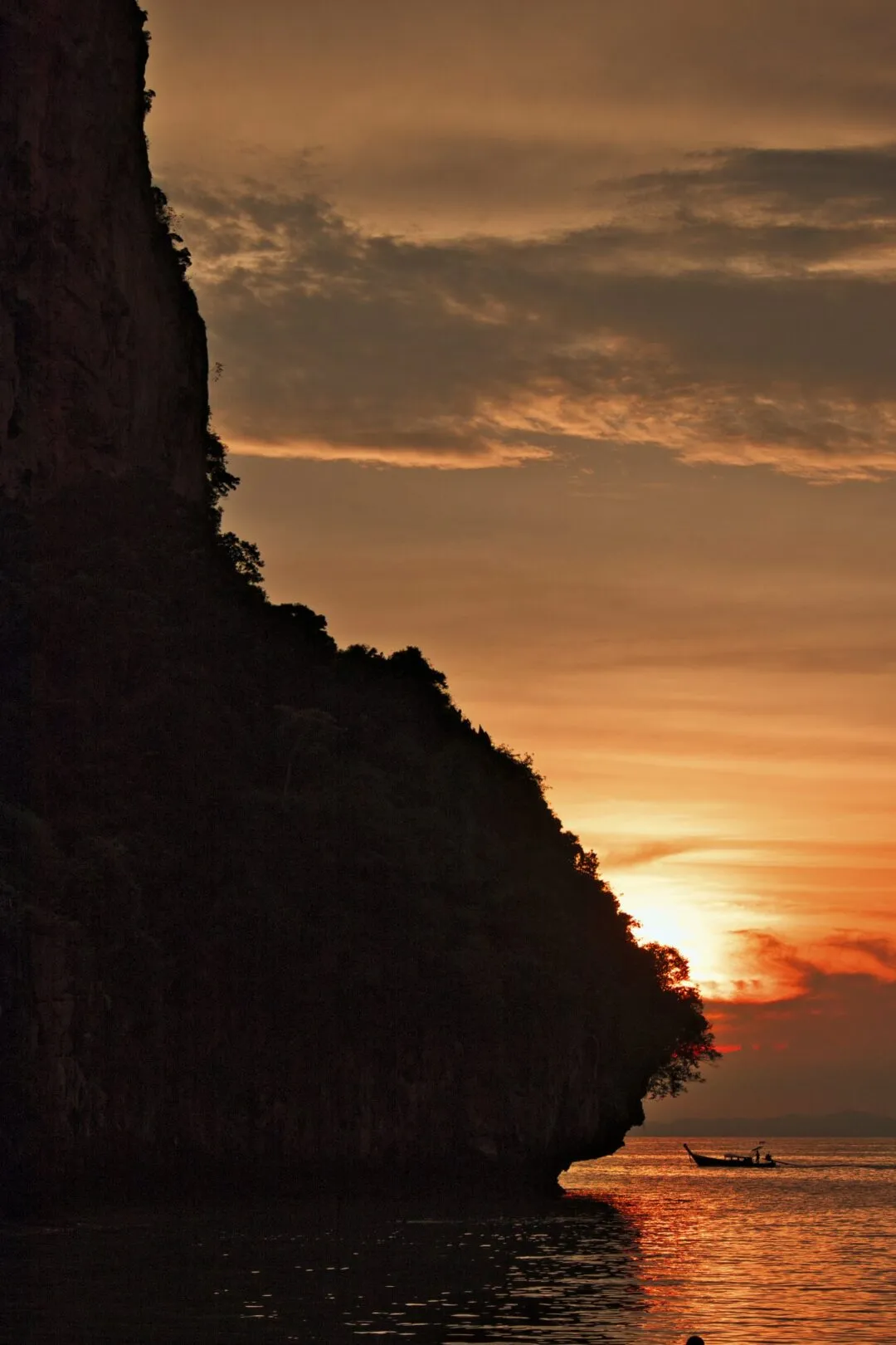 A diagonal limestone cliff silhouette at sunset with a boat's glowing wake, Thailand.