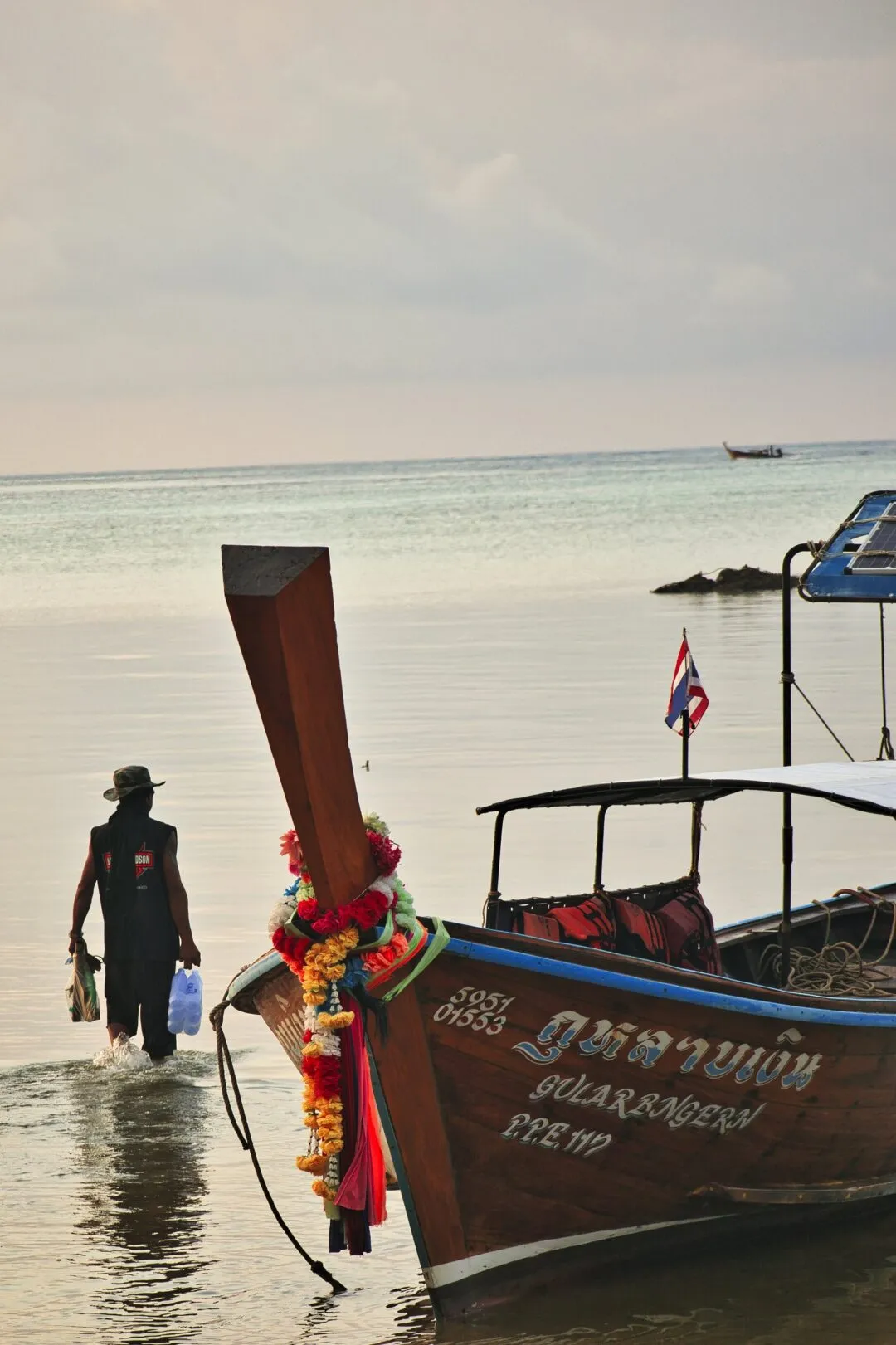 A local fisherman wading ashore beside a garlanded traditional longtail boat in Thailand.