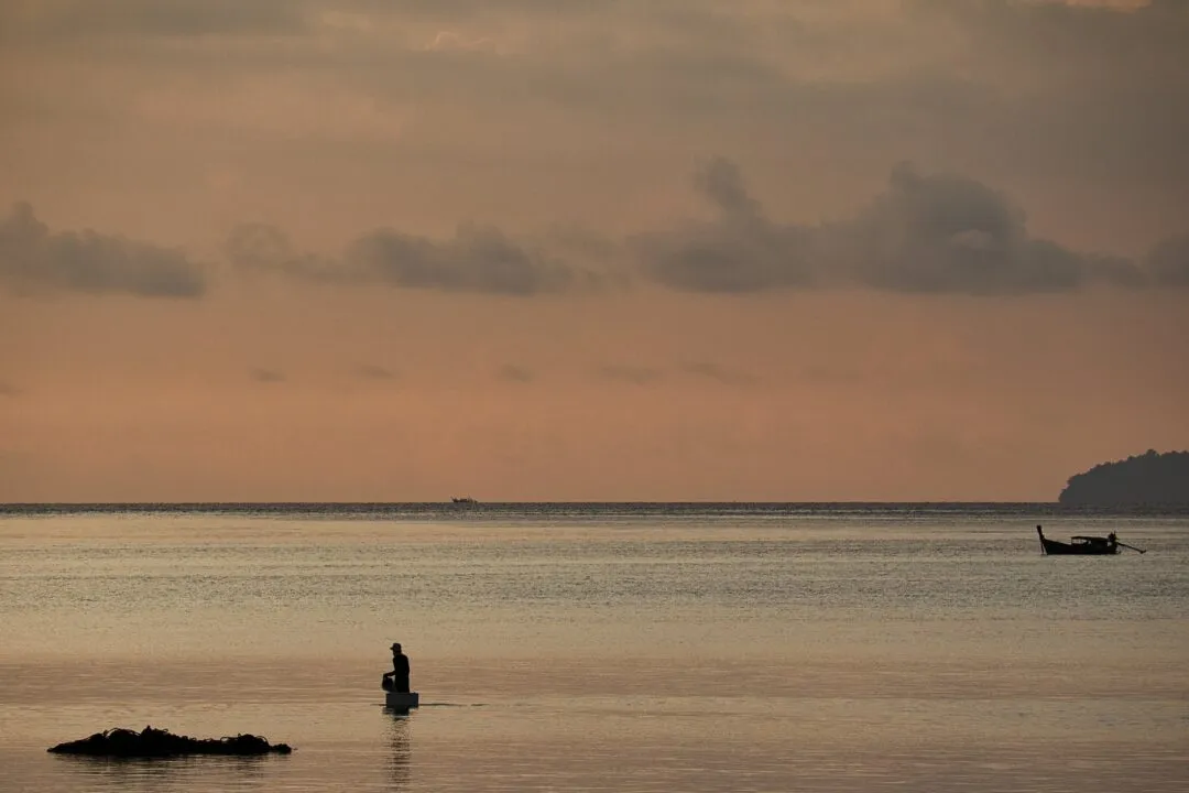 A fisherman silhouetted wading through shallow water at sunset on the Thai coast.