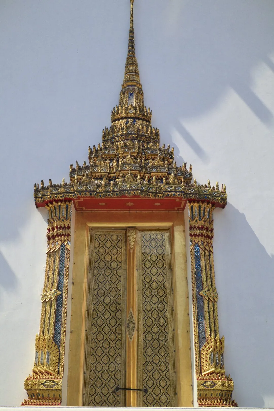 A symmetric gilded temple doorway and spire against a plain white wall in Bangkok.